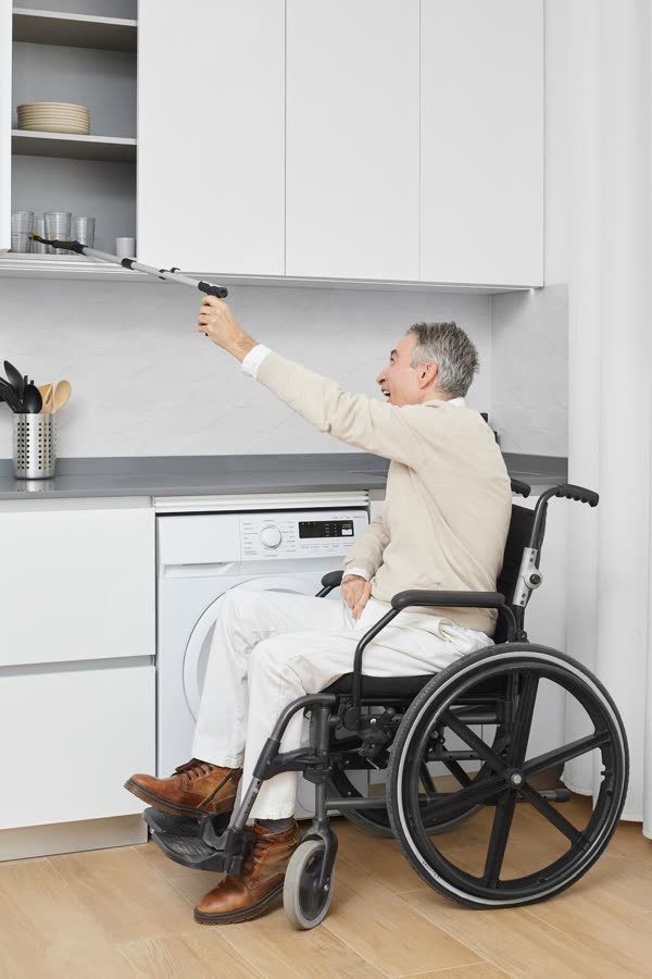 Un hombre en silla de ruedas alcanza un vaso del armario alto de la cocina.